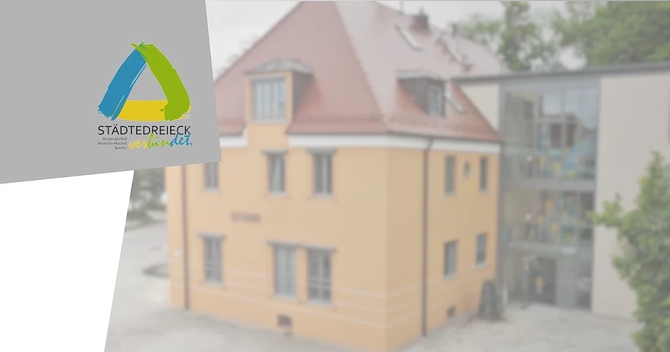 Grafik mit dem Logo des Städtedreiecks auf grauem Hintergrund. Im Hintergrund ist unscharf ein Gebäude mit gelber Fassade und modernem Glasanbau zu sehen. Die Grafik dient als Titelbild für eine Meldung über die Betriebsurlaube der Kleiderkammer im Städtedreieck und des Interkommunalen Recyclinghofs.