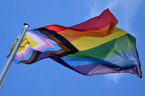 Eine Flagge in Regenbogen- und weiteren Farben weht im Wind vor blauem Himmel.