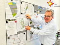 Rüdiger Ropinski steht in seinem Büro an einer voll behangenen Pinnwand und schaut in die Kamera. Er trägt ein Hemd.