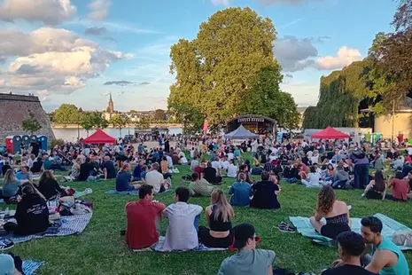 Eine große Gruppe an Personen sitzt im Gras und lauscht einer Band, die auf der Bühne steht
