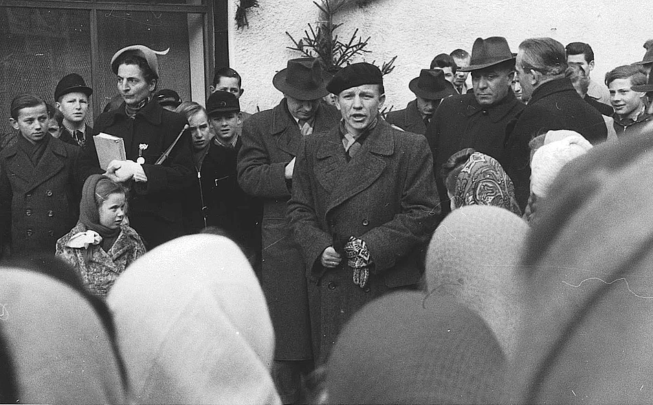 Eine historische Schwarz-Weiß-Aufnahme zeigt einen Redner mit Baskenmütze, der während der Eröffnungsfeier vor einer großen Menschenmenge spricht. Um ihn herum stehen Erwachsene und Jugendliche, während zahlreiche Besucherinnen und Besucher aufmerksam zuhören.