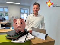 Florian Kapp steht an einer Theke im Vorzimmer im Rathaus. Neben ihm steht ein rosafarbenes Sparschwein. Akten liegen auf der Theke.