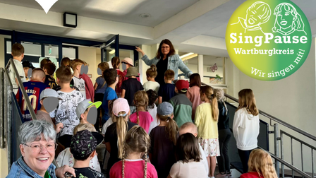 Gruppe von Kindern und Jugendlichen steht auf einer Treppe, eine Frau mit ausgestrecktem Arm spricht zu ihnen, im Vordergrund lächelnde ältere Frau mit Brille und Schal, Logo und Text zu SingPause und Wartburgkreis oben und unten im Bild