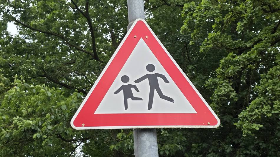 Straßenschild "Achtung Schulkinder" an einem Mast. Im Hintergrund sind grüne Baumblätter.