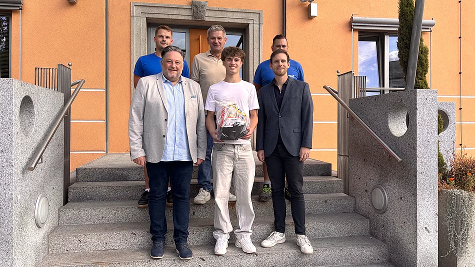 Sechs Männer stehen für ein Gruppenfoto auf der Treppe vor einem orangefarbenen Gebäude. Im Mittelpunkt steht der neue Auszubildende Leonhard Zenger, der einen Geschenkkorb in den Händen hält. Neben und hinter ihm stehen fünf Männer, die ihn zum Beginn seiner Ausbildung willkommen heißen.