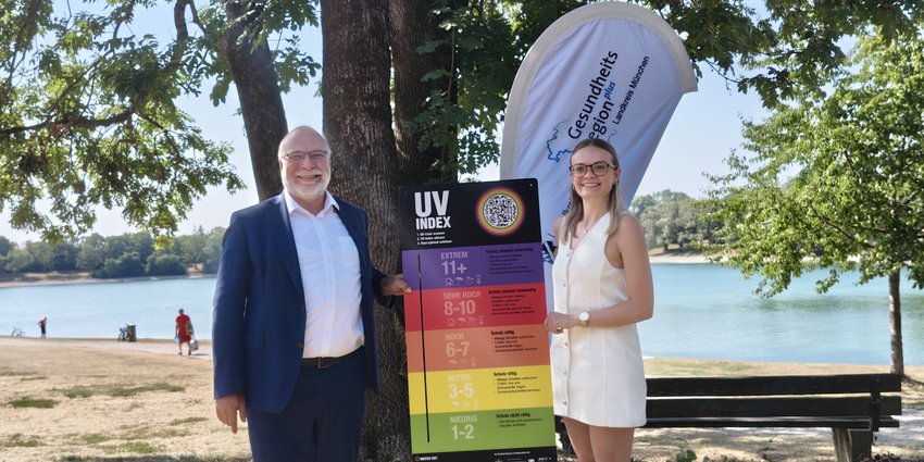 Foto: Ein Mann und eine Frau stehen vor einem Baum an einem See und halten eine Tafel mit mehreren Farben - eine UV-Index-Tafel - in die Kamera