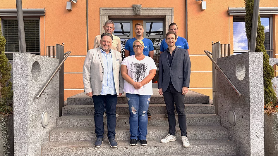 Sechs Personen stehen für ein Gruppenfoto auf der Treppe vor einem orangefarbenen Gebäude. Im Mittelpunkt steht Kathrin Steger. Neben ihr stehen zwei Männer, dahinter drei weitere Männer. Die Gruppe blickt lächelnd in die Kamera und heißt die neue Mitarbeiterin im Mensa-Team willkommen.