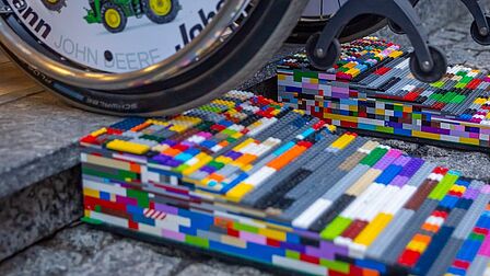 Rollstuhlrad fährt über eine bunte Rampe aus Lego-Steinen auf einem gepflasterten Boden
