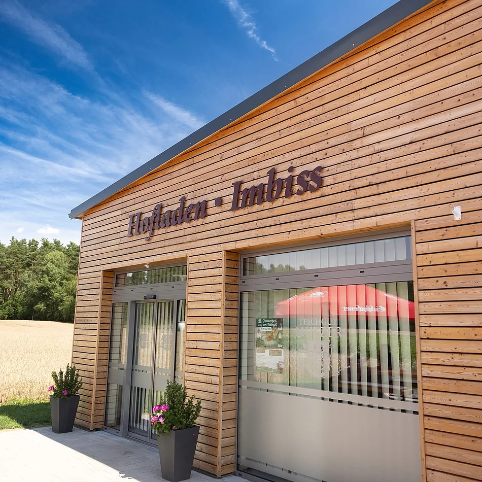 Das Foto zeigt den modernen Holzbau des Hofladens und Imbisses des Teichguts Mulzer mit großen Glasfronten und der gut sichtbaren Aufschrift „Hofladen · Imbiss“. Im Hintergrund sind ein Getreidefeld und ein Waldrand unter blauem Himmel zu erkennen.