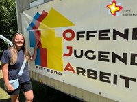 Julia Schmitfranz steht draußen an eine Wand gelehnt, die die Aufschrift Offene Jugend Arbeit trägt.