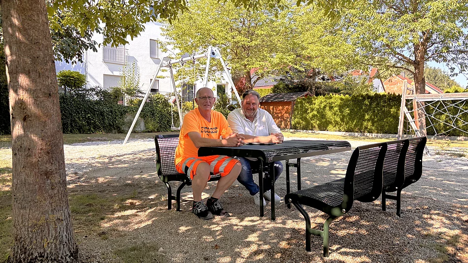 Zwei Männer sitzen an einer neuen Sitzgruppe auf dem Spielplatz „Am Schafgraben“, im Hintergrund sind die erneuerte Doppelschaukel sowie weitere Spielgeräte unter schattenspendenden Bäumen zu sehen.