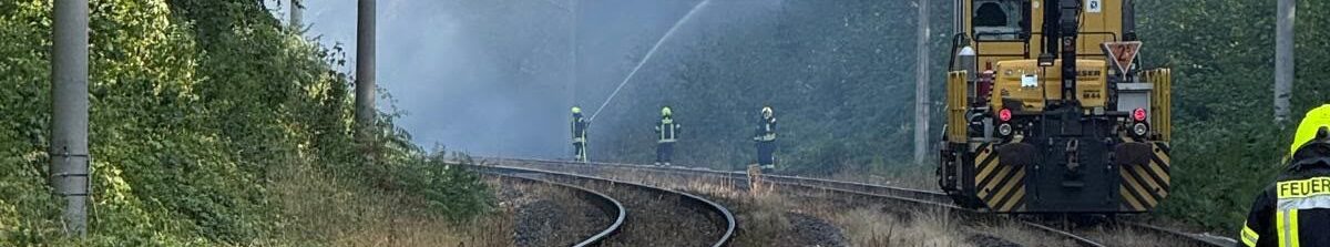 Feuerwehrkräfte löschen einen Böschungsbrand entlang einer Bahnstrecke. Im Hintergrund sind Rauch sowie ein Schienenfahrzeug auf den Gleisen zu sehen.