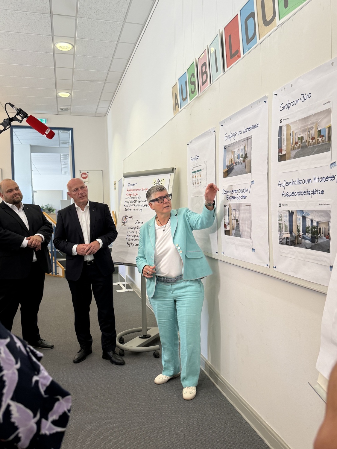Drei Personen stehen vor einer Wand mit Infomaterial und einem Flipchart. Eine Person zeigt auf einen Zettel.