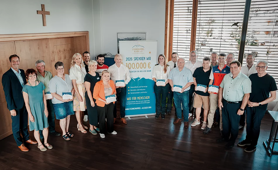 Männer und Frauen stehen in einem Raum für ein Gruppenbild zusammen. Einige von Ihnen halten ein blau/weißes Papier in den Händen und präsentieren es in die Kamera. In der Mitte steht ein Aufsteller.