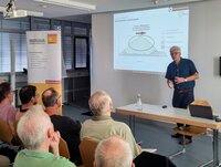 Rudolf Bersch steht an einem Tisch im Sitzungssaal des Rathauses. Hinter ihm ist an der Wand eine Grafik aus seinem Vortrag zu sehen. Vor ihm sitzen Zuschauer und hören ihm zu.
