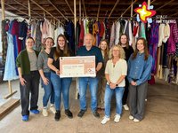 Sieben Frauen und ein Mann stehen - einen übergroßen Scheck haltend - nebeneinander vor einer Stange, auf der viele bunte Kleider hängen.