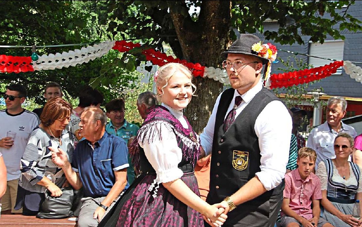 Im Fokus des Bildes ein Tanzpaar in Tanzhaltung. Sie stehen zueinander und halten sich an den Händen. Der Mann trägt ein weißes Hemd dazu eine dunkle Weste, eine dunkle Hose und einen dunklen Hut. An dem Hut steckt eine buntes Blumensträußchen. Seine Tanzpartnerin trägt eine weiße Bluse und ein dunkles Dirndl mit einem lila Tuch und einer lila Schürze. Beide lächeln - im Hintergrund sind Zuschauer zu sehen.