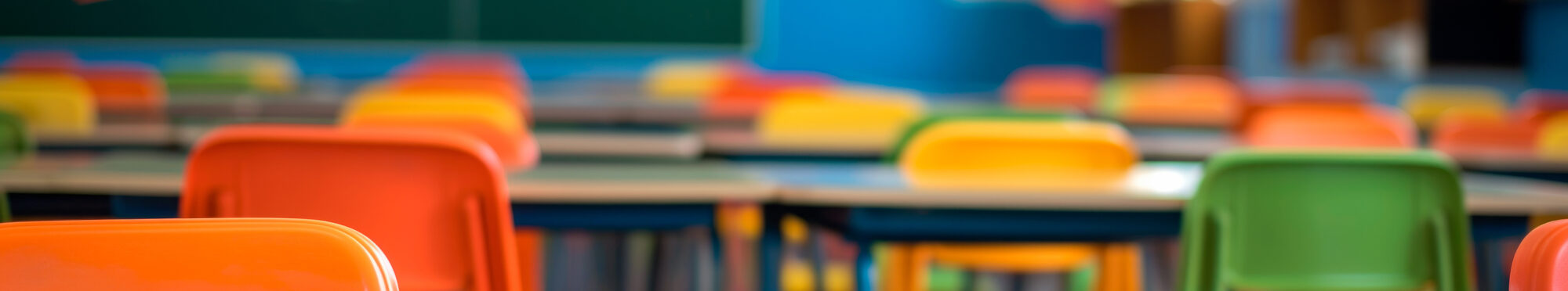 Leeres Klassenzimmer mit mehreren Tischreihen und bunten Stühlen in Orange, Gelb und Grün. Im Hintergrund ist unscharf eine große Tafel zu sehen.