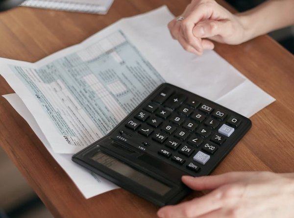 Foto von oben, auf Holzschreibtisch liegt schwarzer Taschenrechner, Formulare liegen darunter, Hand gibt auf Tastatur Zahlen ein. Infos zur Zweitwohnungssteuer