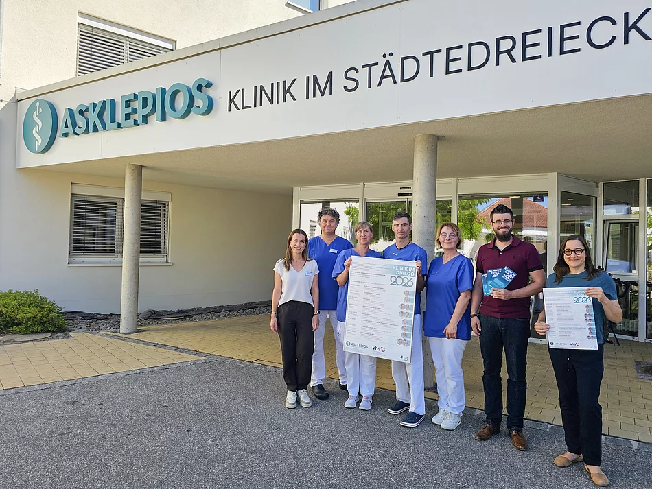 Sieben Personen stehen vor dem Eingang der Asklepios Klinik im Städtedreieck, darunter mehrere Mitarbeitende in medizinischer Berufskleidung; zwei von ihnen halten Plakate zur Veranstaltungsreihe „Klinik im Dialog 2026“ in die Kamera.