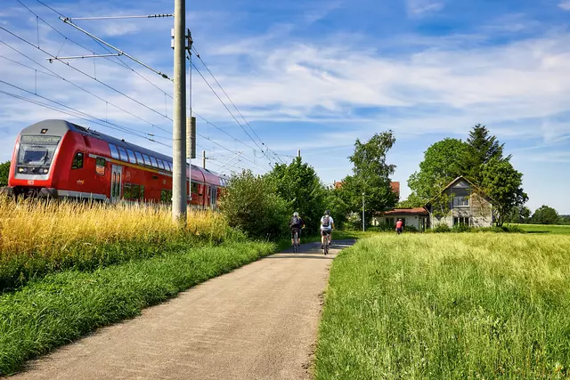Roter Zug neben Radweg in grüner Natur. Im Hintergrund ist ein Bahnwärterhaus zu sehen.