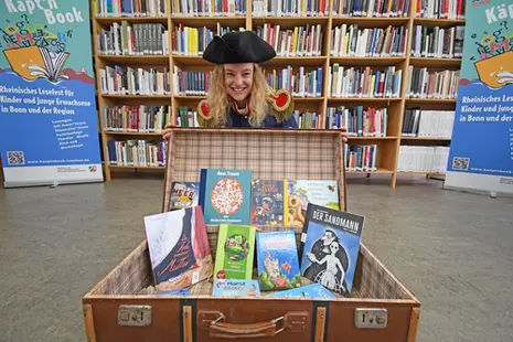 Käpt’n Book - 2021 erstmals von der Schauspielerin Alina Rohde verkörpert -  präsentiert zwischen Bücherregalen einen Koffer voller Bücher