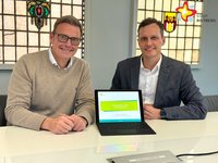 Die genannten Personen sitzen nebeneinander an einem Tisch, schauen in die Kamera. Zwischen ihnen steht ein kleiner Laptop.