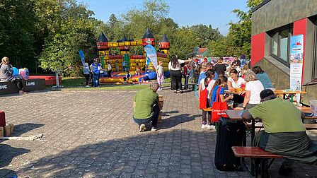 Menschen sitzen und stehen an Tischen im Freien neben einem Gebäude, im Hintergrund eine bunte Hüpfburg und Bäume