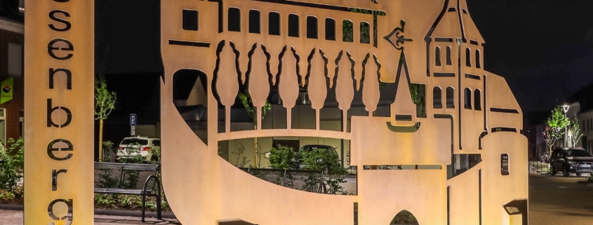 Metallskulptur von Wassenberg mit einem Turm und Bäumen, beleuchtet von unten, auf gepflastertem Weg, bei Nacht.