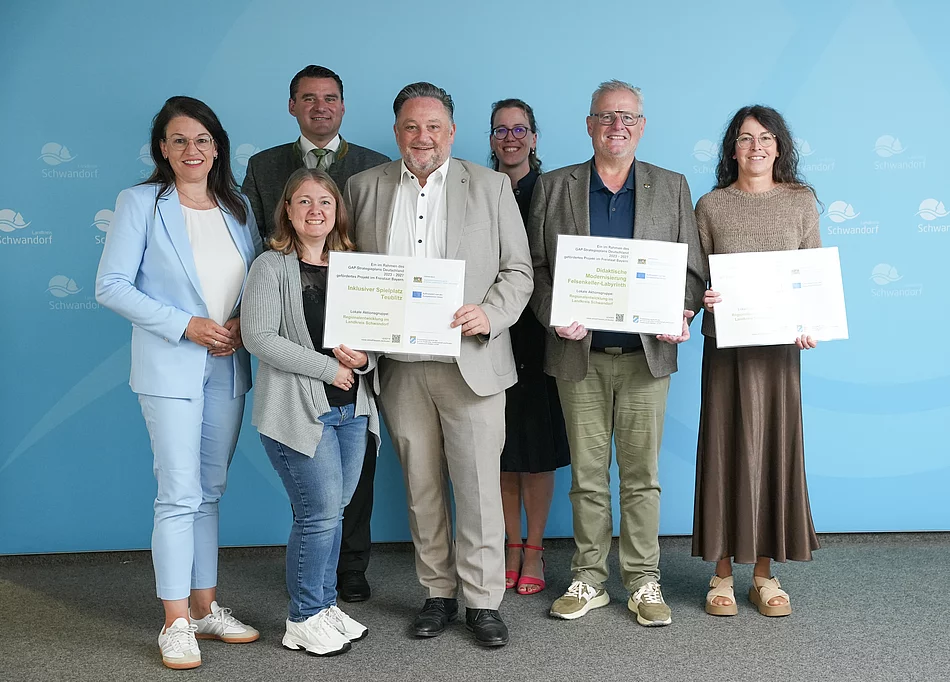 Gruppenfoto von sieben Personen bei der Übergabe mehrerer LEADER-Förderbescheide im Landratsamt Schwandorf. Im Vordergrund halten drei Personen große Fördertafeln in den Händen. Eine davon weist die Förderung für den „Inklusiven Spielplatz Teublitz“ aus. Die Gruppe steht vor einer blauen Wand mit dem Logo des Landkreises Schwandorf.