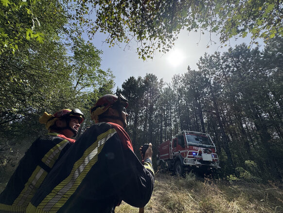Zwei Feuerwehrleute stehen in Vordergrund, im Hintergrund ist ein Feuerwehrauto in einem Waldstück zu sehen.