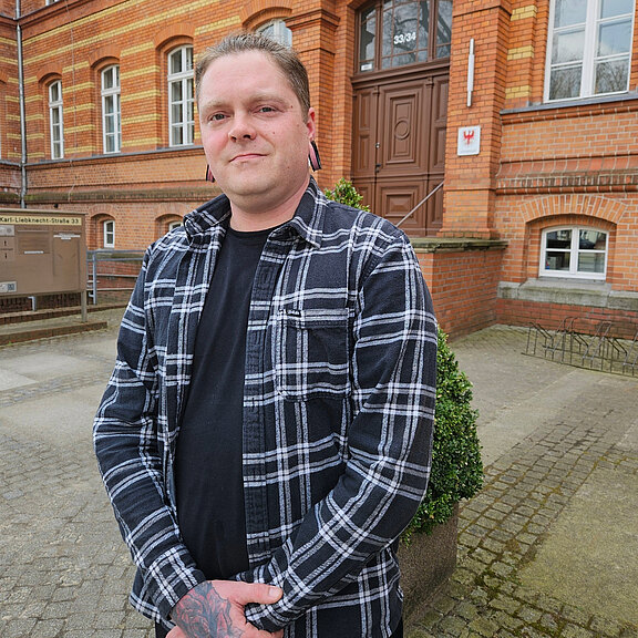 Ein Mann steht vor einem Backsteingebäude, trägt ein kariertes Hemd und hat Tattoos an den Händen. Er blickt direkt in die Kamera und hat eine entspannte Haltung. Im Hintergrund sind Fenster und eine kleine Pflanze sichtbar.
