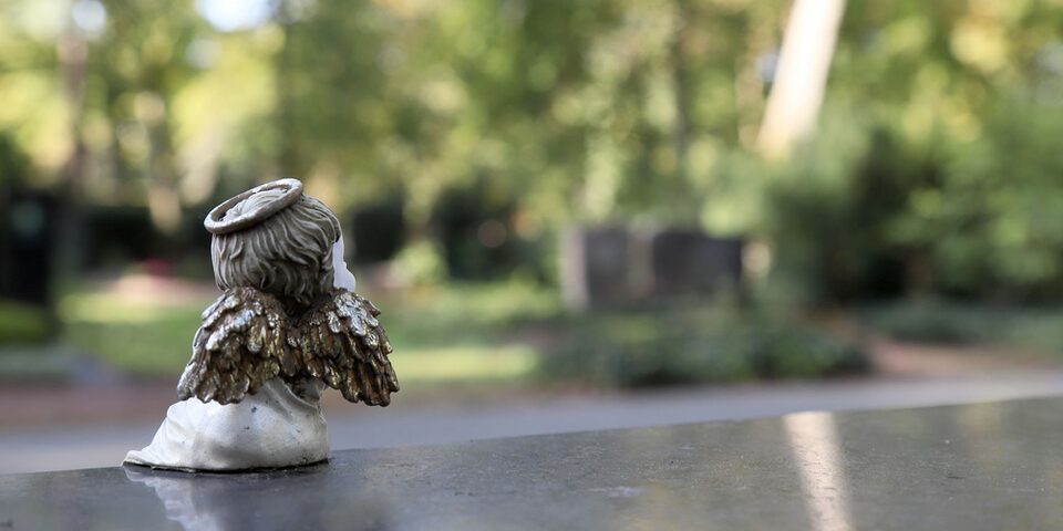 Kleine Engelsfigur steht auf einem Grabstein. Im Hintergrund verschwommene Bäume und Grabsteine.