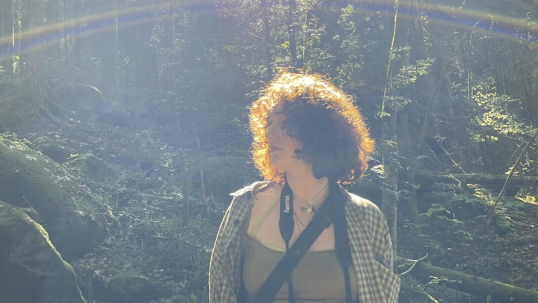 Eine Person steht vor Bäumen. Sie hat eine Kamera um den Hals hängen.