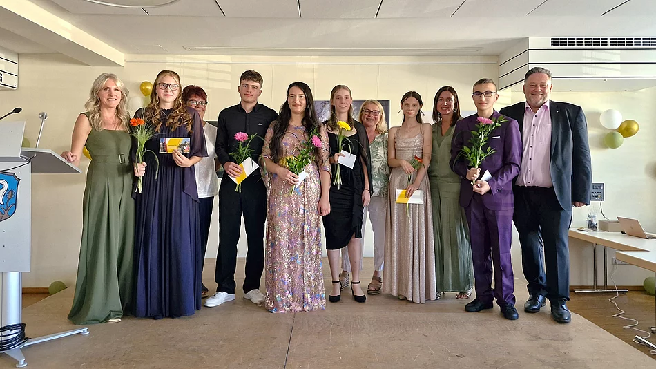 Schülerinnen und Schüler der Abschlussklasse stehen gemeinsam mit ihrer Klassenleiterin, weiteren Lehrkräften und Bürgermeister Thomas Beer auf der Bühne der Abschlussfeier. Die Absolventinnen und Absolventen halten Blumen und ihre Abschlussunterlagen in den Händen und blicken lächelnd in die Kamera.
