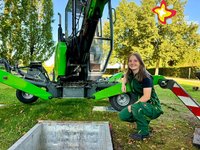 Eine junge Frau in grüner Arbeitskleidung kniet auf einer grünen Wiese hinter einem gerade ausgehobenen Grab und vor einem speziellen Friedhofsbagger