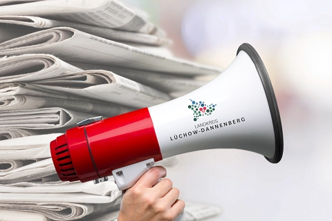 Hand hält ein weiß-rotes Megafon vor einen Stapel Zeitungen mit Logo „LÜCHOW-DANNENBERG“. © Canva