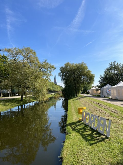 Das Bild zeigt die Königswiesen in Schleswig. Mittig fließt Wasser und im Hintergrund ist der Schleswiger Dom. Rechts im Bild sind einige weiße Zelte des Norden Festivals aufgebaut und davor steht ein Schild bestehend aus den Buchstaben des Wortes "NORDEN"..