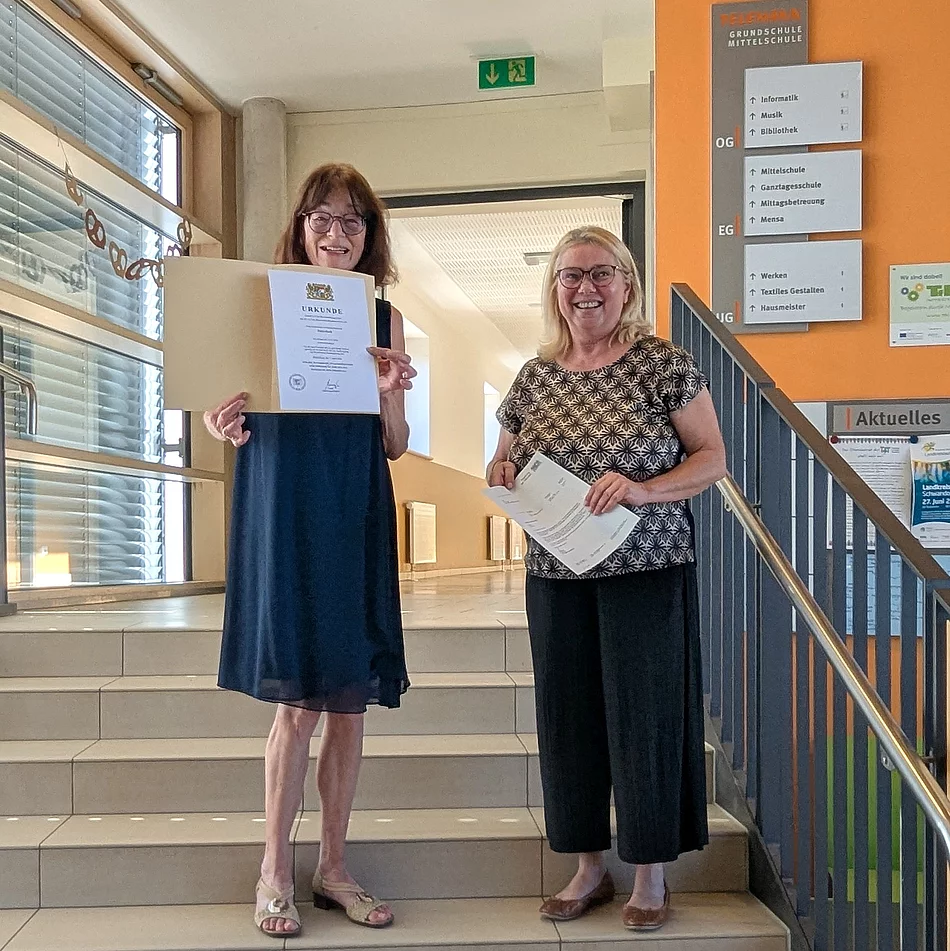 Zwei Frauen stehen lächelnd auf einer Treppe im Eingangsbereich der Telemann-Grundschule. Petra Koch hält ihre Pensionsurkunde in den Händen, daneben steht Schulleiterin Sabine Kunz mit weiteren Unterlagen.
