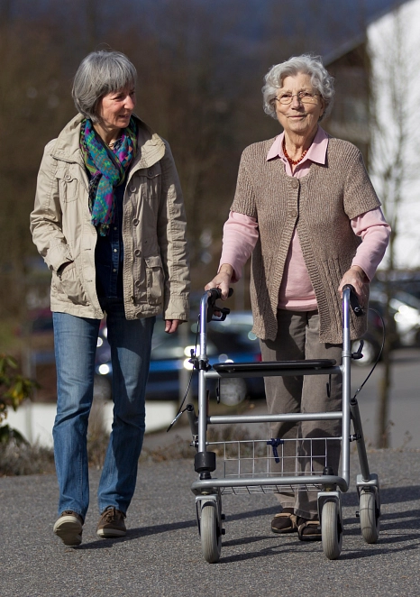 Eine Frau läuft neben einer älteren Frau, die einen Rollator hat. Beide sind in guter Stimmung und Lächeln. Im Hintergrund sieht man Bäume. © Adobe Stock