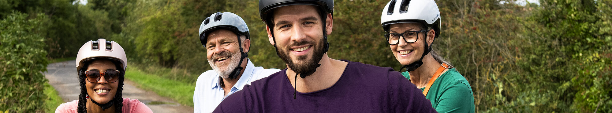 „Vier Erwachsene unterschiedlichen Alters stehen mit Fahrradhelmen und Fahrrädern auf einem Weg im Grünen. Im Vordergrund hält ein Mann ein E-Bike, die Gruppe blickt lächelnd in die Kamera.