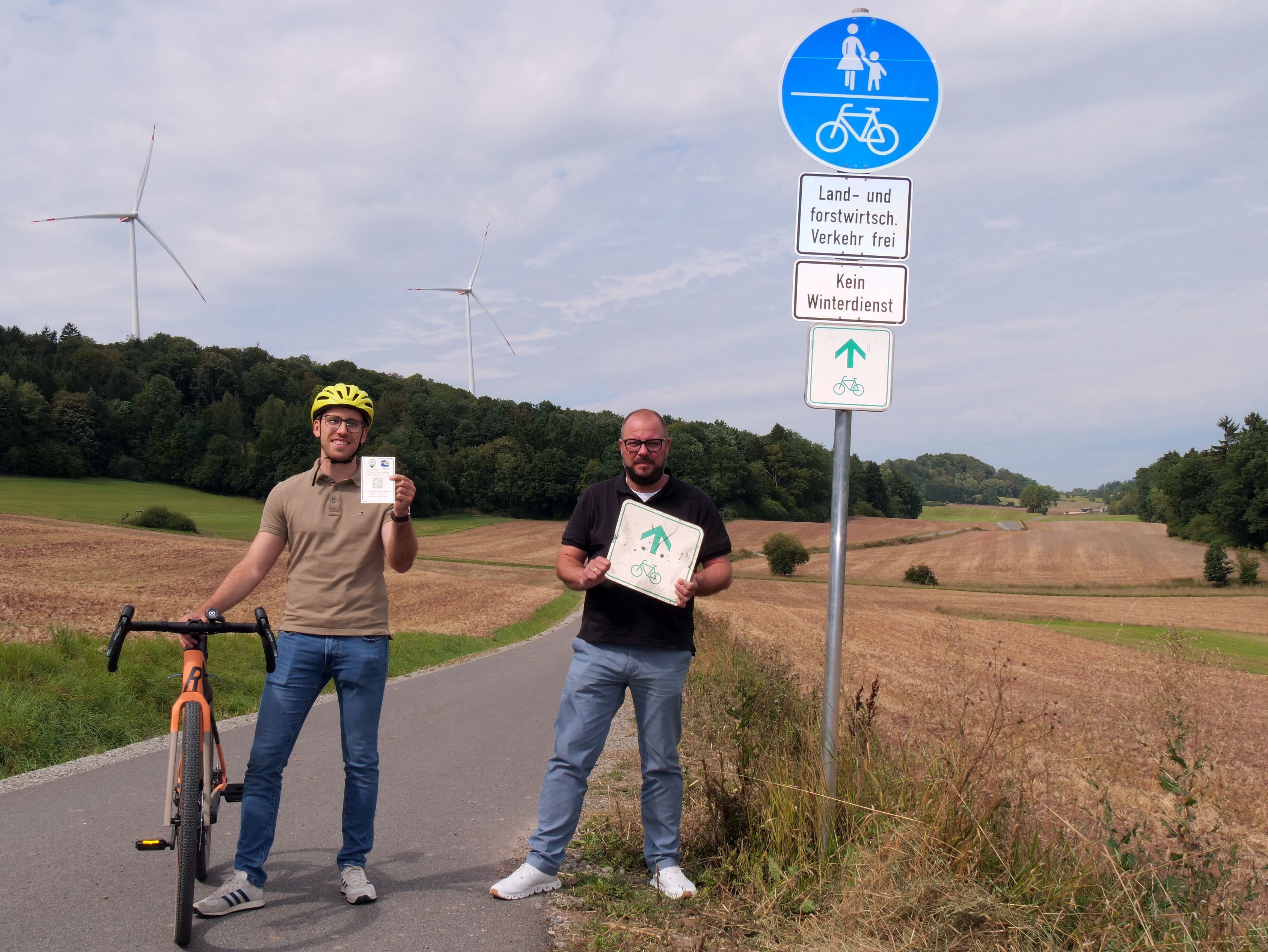 Dominik Wank steht neben seinem Fahrrad und hat einen Aufkleber mit QR-Code in der Hand, neben ihm Sebastian Straubel mit einem unleserlichen Radwege-Schild. Beide stehen auf einem Radweg, im Hintergrund sind zwei große Windräder zu sehen. Damit erkennt man, dass das Bild bei Tremersdorf entstanden ist.
